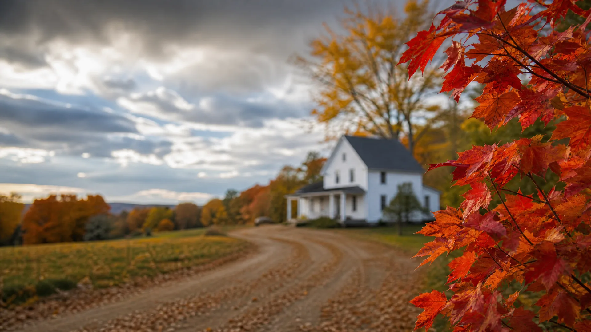 A dirt road curves toward a white two-story farmhouse with a porch, framed by red maple leaves in the foreground under a cloudy sky, with autumn-colored trees and a field alongside.